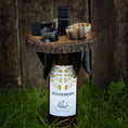 Indlæs billede i Gallery viewer, En flaske Hyldemjød mjød, beliggende delvist i græs, er omgivet af et rustikt Holder til vin med horn med sorte buede horn accenter. Det cirkulære træstativ indeholder små horn til servering af drikkevarer og hviler mod en træoverflade i baggrunden.
