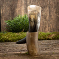 Indlæs billede i Gallery viewer, Drikkehorn Yggdrasil U. Mønster og dens stand, begge fremstillet af poleret hornmateriale, er fremvist på en rustik træoverflade med grønt mos og omkringliggende vegetation. Hornet viser en indviklet indgravering af Yggdrasil, der fremkalder den gamle magi, der minder om et vikingemarked.