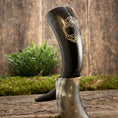 Indlæs billede i Gallery viewer, Drikkehorn Yggdrasil U. Mønster er udstillet opretstående på en træstub, omgivet af grønt mos og planter. Dette elegante drikkehorn har indviklede udskæringer med bladmotiver ætset ind i overfladen, der minder om gamle vikingermærkede skatte og påkalder Yggdrasils ånd.
