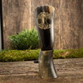 Indlæs billede i Gallery viewer, Et poleret Drikkehorn Yggdrasil U. Mønster er lodret placeret på en rustik træoverflade, med grønt mos og græs i baggrunden mod en struktureret trævæg. Scenen udstråler en naturlig og jordnær stemning, der minder om skatte fundet på et vikingemarked.