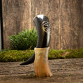 Indlæs billede i Gallery viewer, En poleret Drikkehorn Templar står oprejst i en beige meleret holder på en rustik træoverflade. Frisk grønt løv og mos fremhæver de jordiske omgivelser, og hornet har et indviklet udskåret design nær toppen, der minder om dem, man finder på vikingemarkeder.