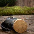 Indlæs billede i Gallery viewer, Et nærbillede af Mini Vikingekrus Horn Thors Hammer (100 ml) hviler på siden på en rustik træoverflade. Hornet, der minder om dem, der vises i Roskilde showroom, er lavet af poleret sort materiale med en lys træbase og har en let sløret naturlig grøn baggrund i baggrunden.