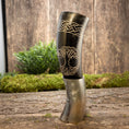 Indlæs billede i Gallery viewer, Et dekorativt Drikkehorn Yggdrasil med Mønster, prydet med udskårne designs, står på en træoverflade. Den øverste halvdel viser indviklede keltiske knudemønstre og et stiliseret ugleansigt. Baggrunden, der fremkalder stemningen af vikingemarkeder, omfatter grønt mos og planter, der forstærker dens rustikke, naturlige charme.