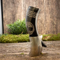 Indlæs billede i Gallery viewer, Drikkehorn Yggdrasil med Mønster, prydet med indviklede hvide udskæringer med et knudedesign og et skjold, hviler på en træoverflade. Bagtæppet fremviser frodigt grønt og en rustik trævæg, der fremkalder atmosfæren af et vikingemarked og forstærker den naturlige og rustikke stemning.