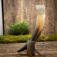 Indlæs billede i Gallery viewer, Drikkehorn Yggdrasil med Mønster, prydet med indviklede keltiske knudedesigns øverst, vises opretstående på en rustik træoverflade ved hjælp af et stativ. Grønt løv og mos i baggrunden skaber naturlige, jordnære omgivelser, der minder om et vikingemarked.