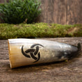 Indlæs billede i Gallery viewer, En Drikkehorn Triscele, prydet med et udskåret triquetra-symbol, ligger på en træoverflade dækket af mos. Grønt løv er let synligt i den slørede baggrund, hvilket skaber en rustik og naturlig indstilling, der minder om vikingemarkeder.