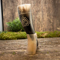 Indlæs billede i Gallery viewer, En håndlavet Drikkehorn Valknut med et slående mønster hviler oprejst på en træoverflade. Hornet har et sort og cremefarvet gradientdesign, prydet med et trekantet emblem. En naturholder støtter den på baggrund af grønne omgivelser og en rustik trævæg.