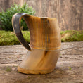 Indlæs billede i Gallery viewer, Et Vikingekrus Bark Horn (600-800ml) med træbund og håndtag hviler på en rustik træoverflade. Hornets krop er poleret med en naturlig gradient af lyse til mørkebrune nuancer, med udskårne lodrette linjer nær toppen. Grønne buske er slørede i baggrunden.
