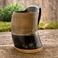 Indlæs billede i Gallery viewer, Et udsøgt håndlavet Vikingekrus Bark Horn (600-800 ml) med en trælignende tekstur på den øverste del og en glat, blank sort underdel, elegant vist på en rustik træoverflade med frodig grønt i baggrunden.