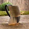 Indlæs billede i Gallery viewer, Et Vikingekrus Bark Horn (600-800ml) lavet af udskåret træ og poleret til en glat finish står på en rustik træoverflade. Hornet har lodrette riller langs kroppen, et sort håndtag og messingaccenter i bunden med grønt mos og træplader i baggrunden.