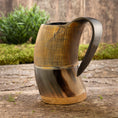 Indlæs billede i Gallery viewer, Et rustikt, håndlavet Vikingekrus Bark Horn (600-800ml) med lodrette kanter på den øverste halvdel og et glat, buet håndtag sidder på en træoverflade. I baggrunden er der grønt og en trævæg, der tilføjer en naturlig, jordnær følelse til scenen.