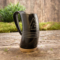 Indlæs billede i Gallery viewer, Et Vikingekrus Valknut Horn (600-800ml), sort og minder om vikingetraditioner, sidder på en træbase på en rustik træoverflade. Hornet er indgraveret med et trekantet design og tre vandrette linjer. I baggrunden er der grønt og en trævæg, der forstærker den naturlige og rustikke stemning.