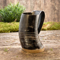 Indlæs billede i Gallery viewer, Et poleret Vikingekrus Valknut Horn (600-800ml) med en træbund er sat på en rustik træoverflade. Baggrunden har grønt og en forvitret trævæg, der tilføjer et naturligt element til scenen. Hornet i vikingestil, der ligner en gammel Krus, har dekorative vandrette riller nær toppen.