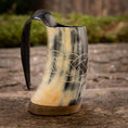 Indlæs billede i Gallery viewer, En Mini Vikingekrus Horn Wave (100 ml) med indviklede keltiske knudeudskæringer og et glat håndtag sidder på en rustik træoverflade. Hornkrusen fremviser en blanding af sorte, hvide og beige nuancer, og dens flade base gør, at den kan stå oprejst.