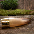 Indlæs billede i Gallery viewer, En Drikkehorn Elegance med indviklet metalarbejde i den brede ende er placeret på en rustik træoverflade. Bagved formidler et lag grønt mos og sløret grønt en naturlig og historisk atmosfære, der minder om vikingemarkeder.