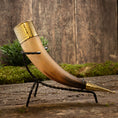 Indlæs billede i Gallery viewer, Drikkehorn Elegance, prydet med indviklede endestykker af guld, hviler yndefuldt i et slankt sort smedejernsstativ. Set mod en rustik trækulisse og omgivet af grønne områder fremhæves hornets håndværk og historiske charme smukt, hvilket gør det til et ideelt stykke for vikingemarkeder.