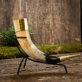 Indlæs billede i Gallery viewer, Drikkehorn Elegance, med et poleret bøffel drikkehorn med en guldudsmykket kant og indviklede detaljer, sidder yndefuldt på et smedejernsstativ. Den rustikke baggrund, prydet med træteksturer og grønt løv, fremkalder stemningen af vikingemarkeder, der forener historisk charme med håndværk.