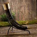 Indlæs billede i Gallery viewer, Drikkehorn Elegance, et dekorativt drikkehorn, holdt i et udsmykket metalstativ prydet med indviklede guldudsmykninger i begge ender, er sat mod en rustik træbaggrund med grønt mos og løv. Denne scene fremkalder vikingemarkedernes vintage charme og en middelalderlig stemning.