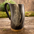 Indlæs billede i Gallery viewer, Et poleret Vikingekrus Valravnen Horn (600-800 ml) har et indgraveret design af en stiliseret fugl på siden. Tankarden viser et sort håndtag og en metallisk base, der hviler elegant på en rustik træoverflade med grønt mos i baggrunden.