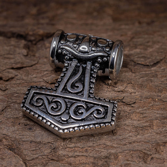 Et nærbillede af Thors Hammer Giant Vedhæng Stål, et sølvvedhæng formet som Thors hammer, Mjölnir, med indviklede detaljer og design, hvilende på en tekstureret træoverflade. Vedhænget har rullemønstre og perler, der viser hammerens form.