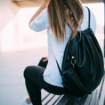 Indlæs billede i Gallery viewer, En person med langt hår og en hvid skjorte sidder på en bænk og vender væk. De bærer sorte jeans og bærer en sort rygsæk med snoretræk med Gede Kaffe Drikkehorn Sort påklipnings BPA-fri vandflaske. Baggrunden er sløret, hvilket antyder en udendørs bymiljø.