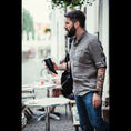 Indlæs billede i Gallery viewer, En skægget mand iført en grå skjorte og jeans står udenfor en café med en Gede Kaffe Drikkehorn Sort. Han bærer en sort skuldertaske og har et tatoveringsærme på venstre arm. Der er tomme borde og stole omkring ham, med slørede individer i baggrunden.