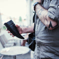 Indlæs billede i Gallery viewer, En person med tatoverede arme, iført en grå skjorte, holder en Goat Kaffe Drikkehorn Sort lavet af BPA-fri plast. De står udendørs nær et rundt bord med en sløret urban baggrund. En skuldertaskerem er synlig på deres højre skulder.