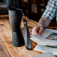 Indlæs billede i Gallery viewer, En person i en ternet skjorte holder en avis med den ene hånd, mens en sort ged Kaffe Drikkehorn Sort lavet af BPA-fri plast og en læder nøglering med teksten "GOAT STORY" er placeret på bordet foran dem. Baggrunden er lidt sløret.