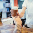 Indlæs billede i Gallery viewer, En tatoveret arm rækker en hånd ud for at tage imod en ged Kaffe Drikkehorn Brun fra en barista på tværs af cafedisken, der minder om en moderne viking, der handler med sit drikkehorn. Baggrunden er sløret og fokuserer scenen på kaffeoverdragelsen.