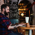 Indlæs billede i Gallery viewer, En skægget mand med tatoveringer, der ligner en moderne viking, er iført en rød og blå plaid skjorte, mens han taster på en bærbar computer ved et rundt træbord i en hyggelig café med bogreoler og tavlevægge. En ged Kaffe Drikkehorn Brun isoleret kop lavet af BPA-fri plastik sidder tæt på hans bærbare computer.
