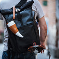 Indlæs billede i Gallery viewer, En person iført en grå t-shirt bærer en stor sort roll-top rygsæk med en hvid og solbrun ged Kaffe Drikkehorn Brun påsat, mens han skubber en cykel med en brun Brooks-sadel i læder. Scenen fremkalder stemningen fra en moderne viking på en bygade.