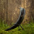 Indlæs billede i Gallery viewer, Drikkehorn Helleristning, der byder på kunstfærdige udskårne designs og en poleret finish, er elegant udstillet på et metalstativ. Den ligger på græs, med et plankeværk i baggrunden, hvilket skaber en rustik og historisk atmosfære, der minder om vikingemarkeder.
