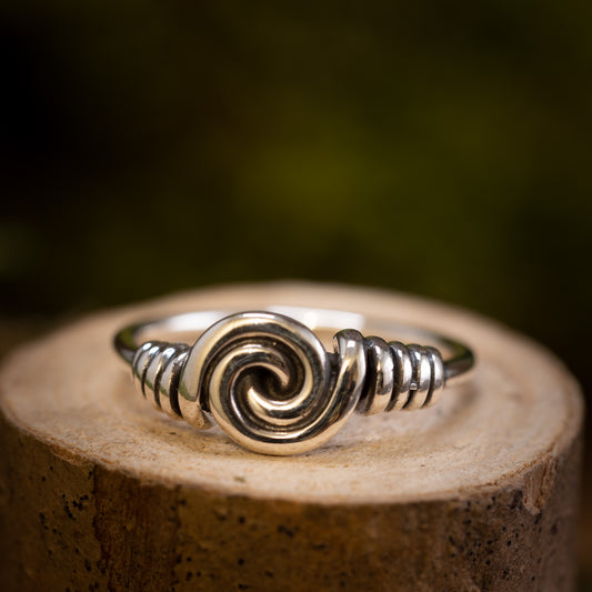 Sølvring Spiral 925s Sølv er elegant placeret på en træoverflade. Denne sølvring kan prale af et slående spiraldesign i midten, med indviklet wirework, der fremhæver spiralen, der afspejler vikingernes håndværk. Baggrunden er blødt sløret med jordnære og grønne nuancer.