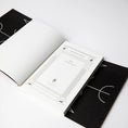 Indlæs billede i Gallery viewer, En hardcover bog er åben for at afsløre en indre hvid forside med titlen "Square's Circle" med dekorative kanter. Det ydre sorte omslag, inspireret af nordiske besværgelser, har et geometrisk design med linjer og delcirkler. Dette produkt, kaldet Sorcerer's Screed - Runestave, ligger på en hvid overflade.
