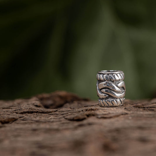 Et nærbillede af Skægperler / Hårpynt Waves Sølv-ringen med et indviklet, vævet slangedesign i Vikingesmykker-stil. 925s Sterling Sølv-ringen er sat på en tekstureret træoverflade med en sløret grøn baggrund.