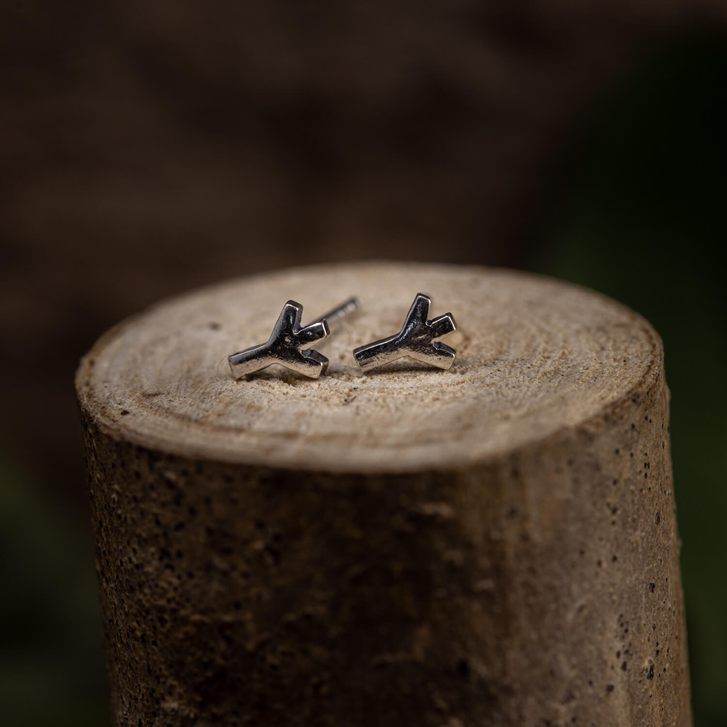 Et par grenformede øreringe, lavet af ægte 925s Sterling sølv, hviler på en træoverflade. Det minimalistiske, naturinspirerede design har en glat, poleret finish. Baggrunden er sløret, hvilket fremhæver træets tekstur og de fine detaljer i disse Ørestikker Runer 925s Sølv.