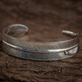 Indlæs billede i Gallery viewer, Et nærbillede af et indviklet udformet sølvmanchetarmbånd. Armbånd Bangle Feather 925s Sterling Sølv har detaljerede, ætsede diagonale linjer, der efterligner teksturen af en kragefjer med en poleret finish. Armbåndet er lavet af 925s Sterling Sølv og er sat mod en rustik, brun træbaggrund.
