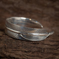 Indlæs billede i Gallery viewer, Et nærbillede af Armbånd Bangle Feather 925s Sterling Sølv designet til at ligne en fuglefjer. Armbåndet har indviklede detaljer, med fine linjer, der efterligner teksturen af en ægte kragefjer. Det er placeret på en rustik træ baggrund.
