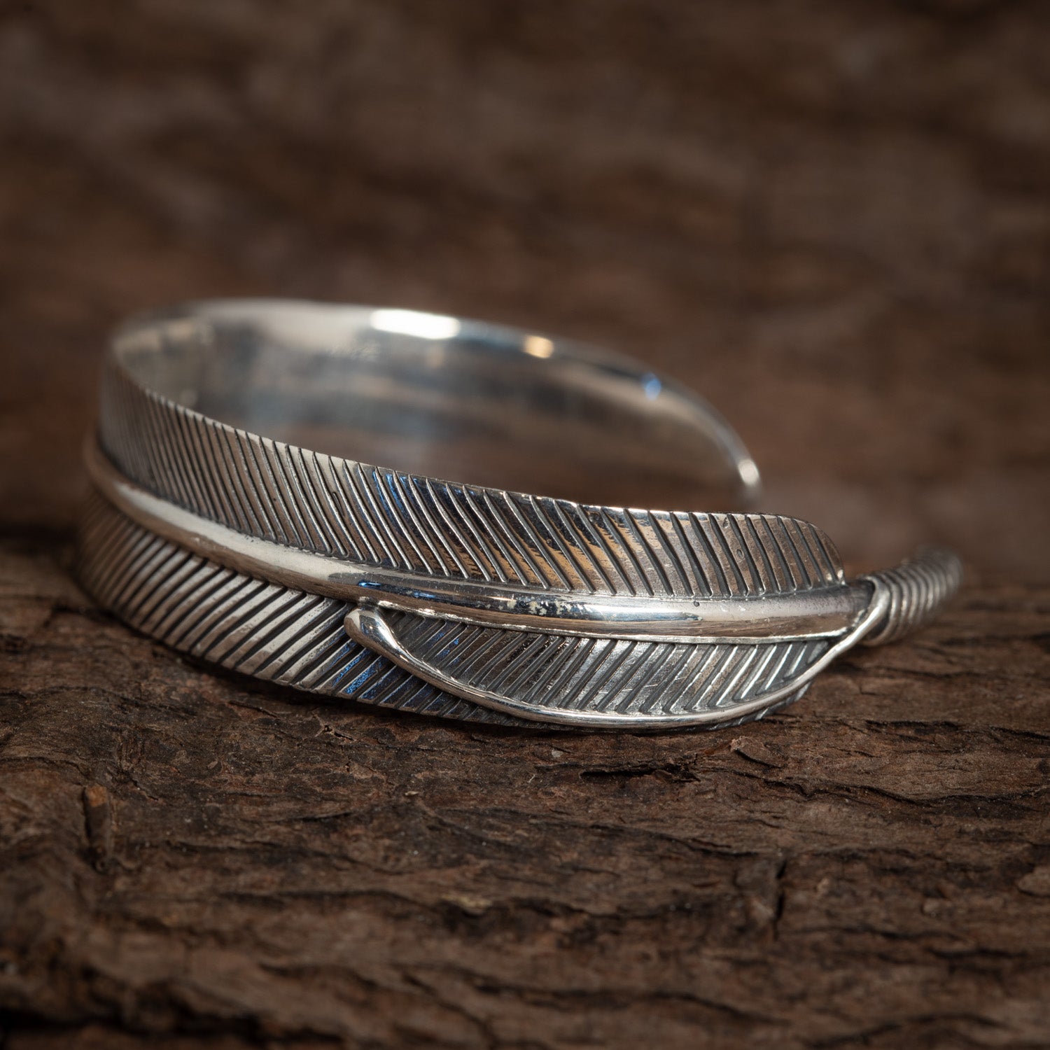 Et nærbillede af Armbånd Bangle Feather 925s Sterling Sølv designet til at ligne en fuglefjer. Armbåndet har indviklede detaljer, med fine linjer, der efterligner teksturen af en ægte kragefjer. Det er placeret på en rustik træ baggrund.