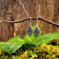 Indlæs billede i Gallery viewer, Et par elegante Hængeøreringe Braid 925s Sølv med indviklede designs og cabochon zirkon ametyst ædelsten hænger fra en rustik trægren. Under dem skaber store grønne blade og mos et levende bunddække, der forstærker de naturlige omgivelser med en skovstemning.
