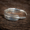 Indlæs billede i Gallery viewer, "Armbånd Bangle Feather 925s Sterling Sølv" er et 925s sterlingsølv armbånd, der er indviklet designet til at ligne en detaljeret kragefjer, med den ene ende snoet rundt for at danne det åbne armbånd, smukt hvilende på en tekstureret træoverflade.
