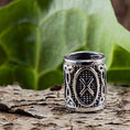 Indlæs billede i Gallery viewer, Et nærbillede af en detaljeret Vikingesmykker-ring med indviklede mønstre og et centralt "X"-design, der minder om Rune Gebo. Skægperler / Hårpynt Rune G Gebo 925s Sølv ringen er placeret på en struktureret, barklignende overflade med et stort grønt blad i baggrunden.
