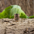 Indlæs billede i Gallery viewer, En omhyggeligt udformet ring, med det indviklede design af Skægperler / Hårpynt Rune J Jera Bronze, hviler yndefuldt på et stykke træbark. Et stort grønt blad i baggrunden fremhæver dets udsøgte geometriske former og delikate udskæringer, der minder om vikingesmykker, hvilket skaber en slående kontrast mod de naturlige, rustikke omgivelser.
