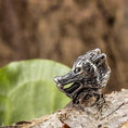 Indlæs billede i Gallery viewer, En Fenrisulv Stål hårpynt, stylet som et ulvehoved, hviler på et stykke bark. Baggrunden er let sløret, med et grønt blad og en struktureret, brun overflade.
