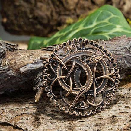 Broche Pitney Spænde Bronze, en autentisk bronze museumskopi, viser indviklede keltisk-inspirerede designs med detaljerede sammenvævede mønstre. Afbildet mod ru træbark og grønne blade i baggrunden har brochen en blanding af løkker og knuder inden for en cirkulær ramme, der minder om Urnestil-perioden.