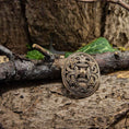 Indlæs billede i Gallery viewer, En bronze Broche Borrestil Bronze hviler på en barkdækket skovbund omgivet af grene og grønt løv. Brochen har indviklede, sammenflettede mønstre, der afspejler gammelt håndværk. Scenen fremkalder en følelse af historie og naturens omfavnelse, der minder om autentisk vikingedesign.
