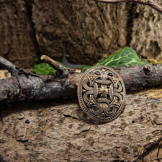 En bronze Broche Borrestil Bronze hviler på en barkdækket skovbund omgivet af grene og grønt løv. Brochen har indviklede, sammenflettede mønstre, der afspejler gammelt håndværk. Scenen fremkalder en følelse af historie og naturens omfavnelse, der minder om autentisk vikingedesign.