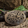 Indlæs billede i Gallery viewer, Et Broche Jellingstil-bæltespænde i bronze med et indviklet keltisk knudedesign med løkker hviler på en tekstureret træbark-baggrund med grønne blade delvist synlige i baggrunden. Metalspændet har et detaljeret, antikt look, der minder om autentisk bronze og afspejler den elegante Jellinge-stilen.
