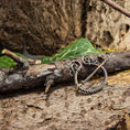 Indlæs billede i Gallery viewer, En Broche Ringnål Antik Bronze, prydet med indviklede løkker og et snoet ringdesign, der minder om Vikingetiden, er fastgjort til en forvitret trægren. Baggrunden har grønne blade og den teksturerede bark af et træ, hvilket skaber en naturlig, rustik indstilling.
