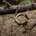 Indlæs billede i Gallery viewer, Broche Ringnål Stor Bronze, prydet med indviklede keltiske designs, hviler på et tekstureret stykke træ. Med trægrene og grønne blade i baggrunden fremkalder denne bronzering minder om vikingegenstande med sine detaljerede graveringer, der har hjertelignende former og spiraler.
